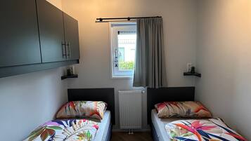 Une chambre avec deux lits, une fenêtre avec des rideaux, un radiateur et un luminaire au plafond.