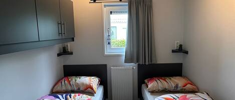 A room with two beds, a window with curtains, a radiator, and a ceiling light fixture.
