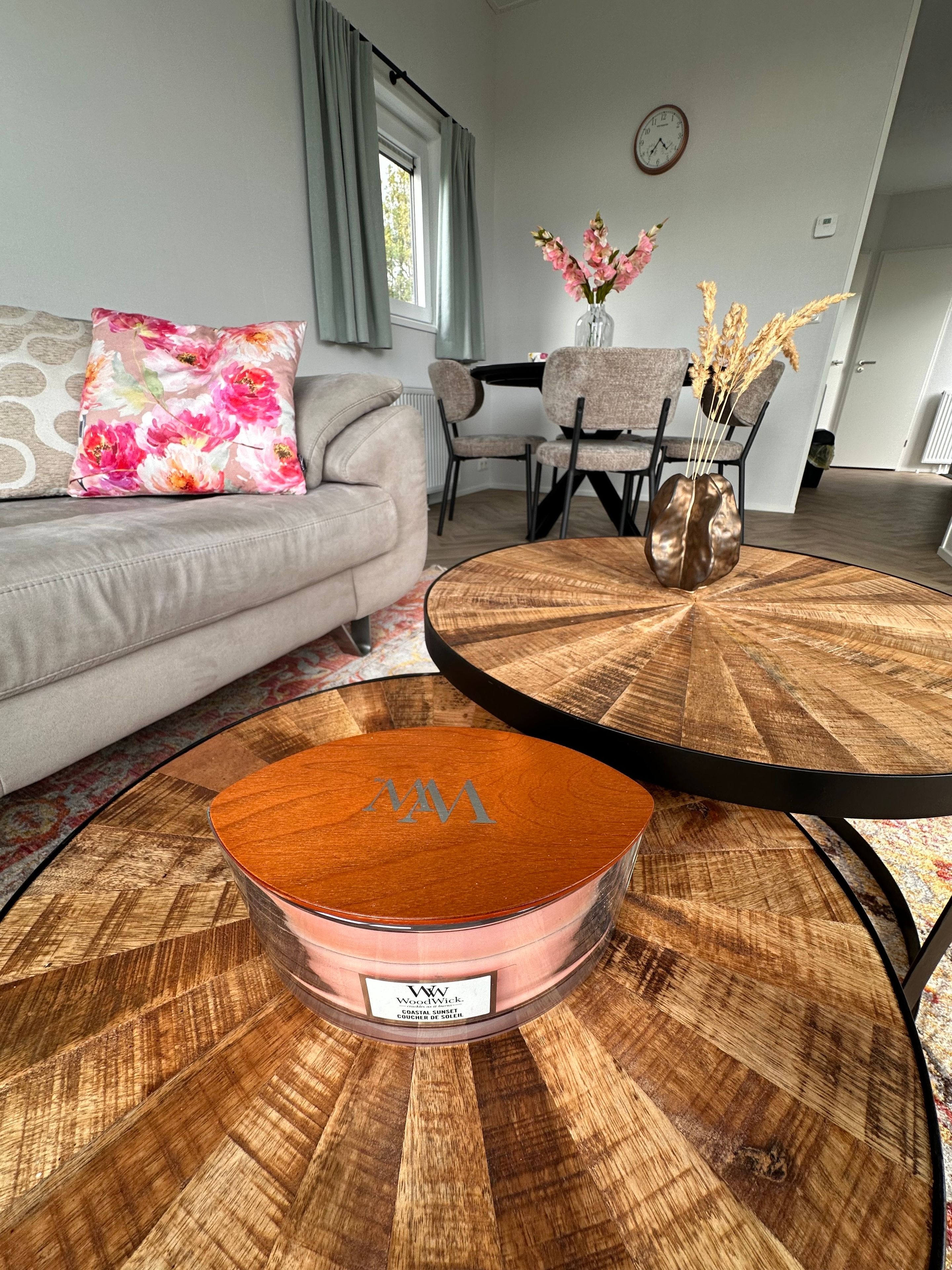Een woonkamer met een ronde houten salontafel, een bank met een bloemenkussen, en een eethoek met stoelen en een bloevas.