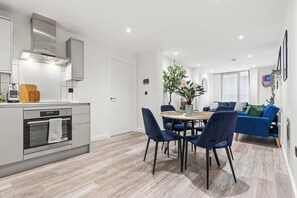 A modern kitchen with a built-in oven, a dining area with a round table and chairs, and a living room with a blue sofa and a plant. - #20 LuxDigbeth Apt|near Hippodrome Square|Sleeps 6 (Deritend)