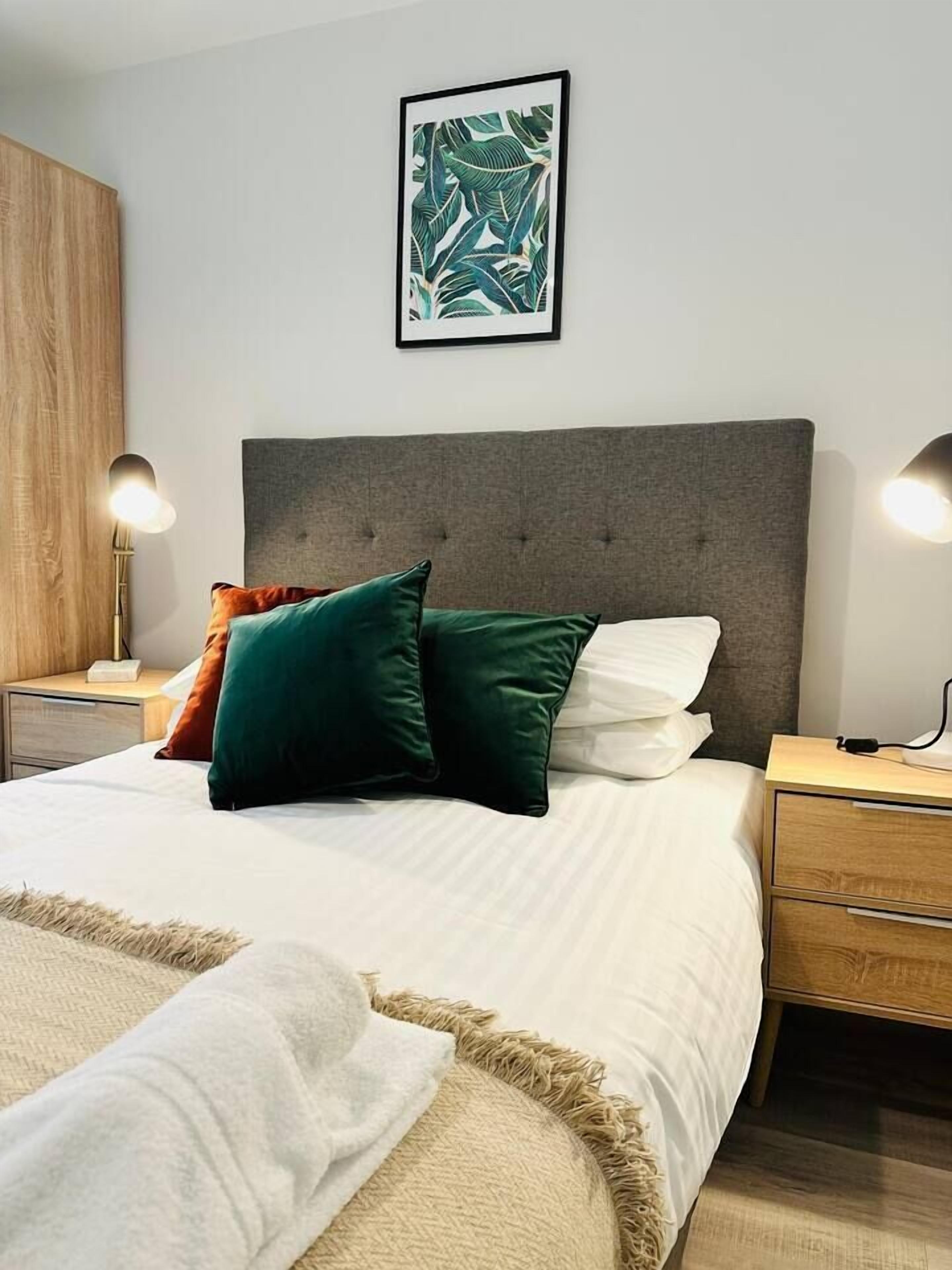 A bedroom with a bed, pillows, bedside tables, a nightstand, a wall-mounted lamp, and a framed picture.