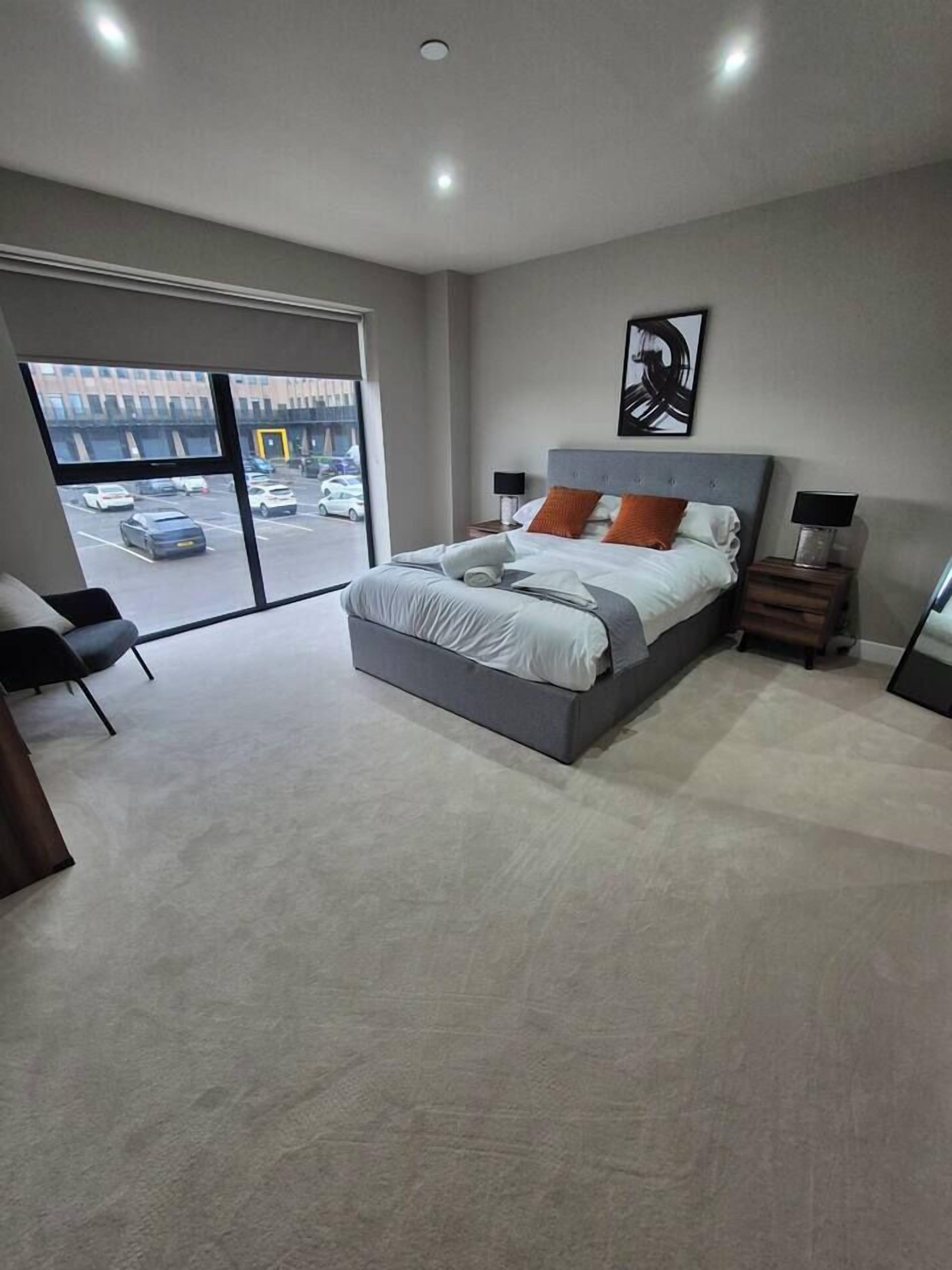 A modern bedroom with a large bed, a chair, a desk, and a view of the parking lot through a sliding door.
