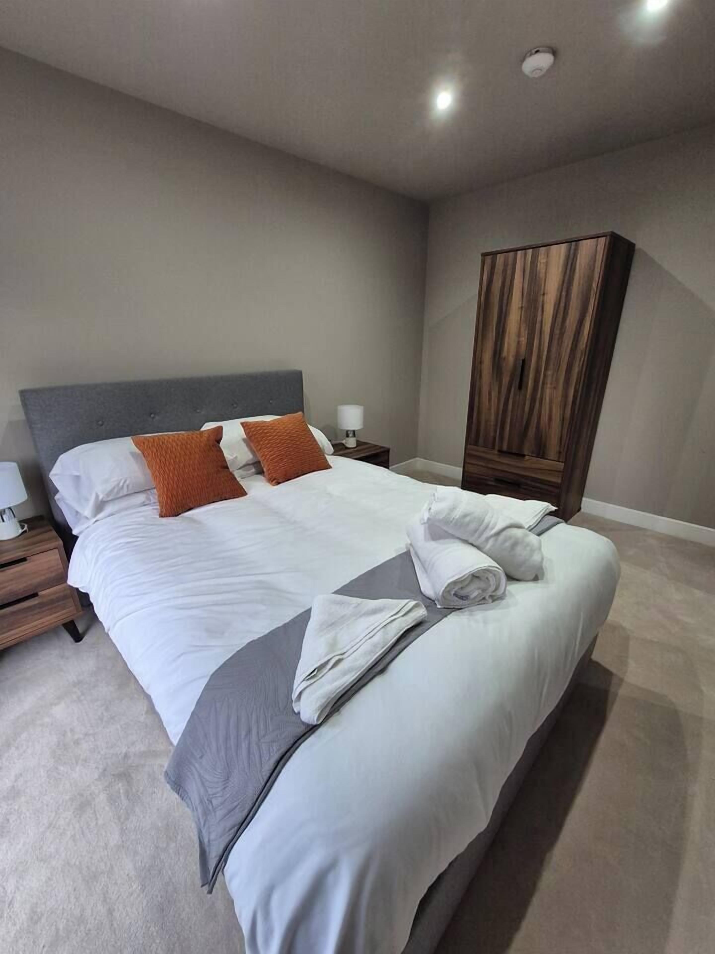 Un lit bien fait, avec du linge de lit blanc et un plaid gris, une armoire en bois et une table de chevet avec une lampe.
