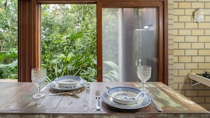 Mesa lista para comer con platos, cubiertos y vasos, frente a una ventana con vistas a vegetación.