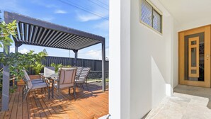 A wooden deck with outdoor furniture and a pergola, a white exterior wall, and a wooden door. - Zuri Luxury Coastal Staycation• 2 Min to Beach, Cafés & Lakefront (The Entrance)