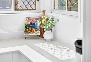A kitchen with a white countertop, a wooden cutting board, a white vase with green plants, and a cookbook titled 'Wholesome' by Sarah Pound. - Zuri Luxury Coastal Staycation• 2 Min to Beach, Cafés & Lakefront (The Entrance)