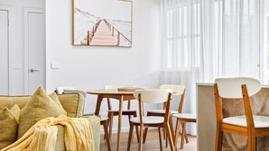 A modern living room with a wooden floor, a sofa with cushions, a coffee table, and a dining area with chairs and a table. - Zuri Luxury Coastal Staycation• 2 Min to Beach, Cafés & Lakefront (The Entrance)