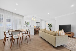 A modern living room with a sofa, TV, and dining area with chairs and a table. - Zuri Luxury Coastal Staycation• 2 Min to Beach, Cafés & Lakefront (The Entrance)