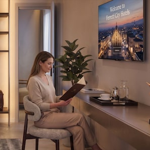 A woman seated in a modern hotel lobby, holding a booklet, with a cityscape view on the wall behind her. - Ferretti Suite Hotel Al Duomo (Milan)