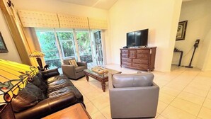 Ein Wohnzimmer mit einem Ledersofa, einem grauen Sessel, einem Holztisch, einem Fernseher auf einer Holzkommode und einem großen Fenster mit Vorhängen.