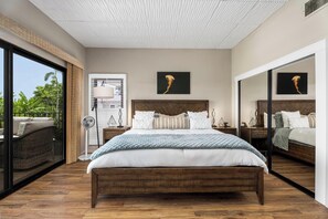 A bedroom with a large bed, a nightstand, a floor lamp, and a sliding glass door leading to an outdoor area. - Honu Family Retreat Royal Seacliff#401 (Kailua Kona)