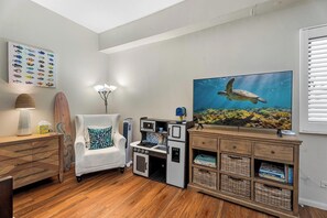 Ein Zimmer mit Holzboden, einem weißen Sessel, einer Kaffeemaschine, einem Surfbrett, einem Gemälde einer Meeresschildkröte und einem an der Wand befestigten Bücherregal.