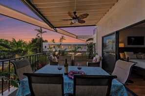 Ein Balkon mit Tisch für zwei Personen, Stühlen, einem Deckenventilator und Blick auf ein Gebäude und Palmen.