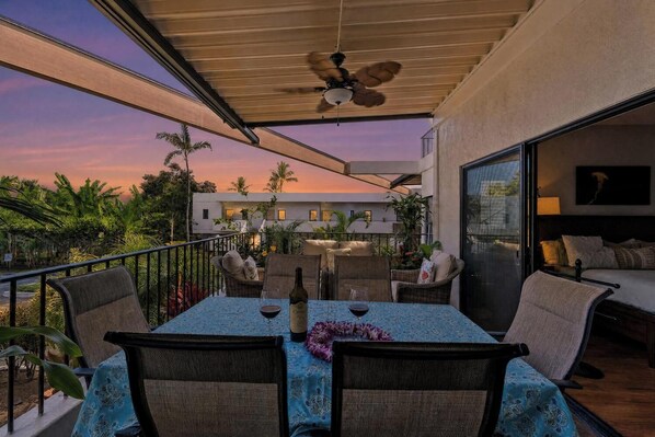 A balcony with a table set for two, chairs, a ceiling fan, and a view of a building and palm trees. - Honu Family Retreat Royal Seacliff#401 (Kailua Kona)