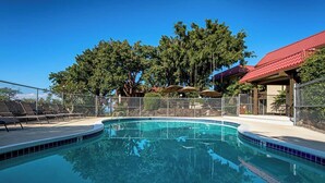 A clear swimming pool with a fenced deck area, surrounded by trees and a building with a red roof. - Kona Mansions#B116 Mountain views, No Stairs, Heart of Town (Kailua Kona)