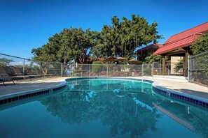 A clear swimming pool with a fenced deck area, surrounded by trees and a building with a red roof. - Kona Mansions#B116 Mountain views, No Stairs, Heart of Town (Kailua Kona)