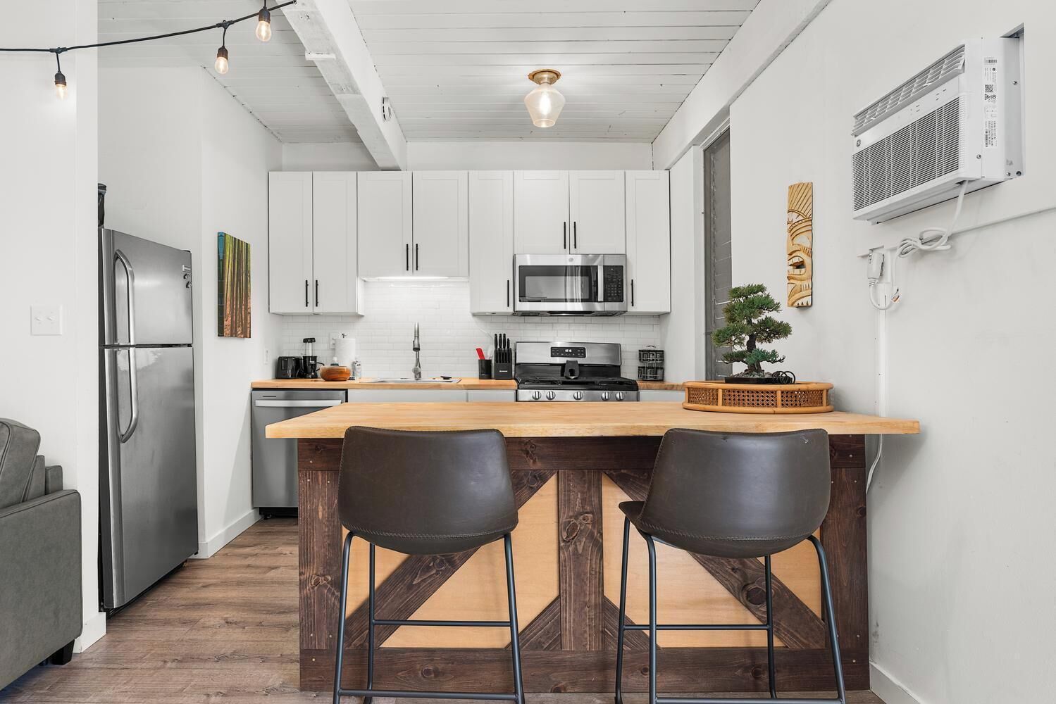 A modern kitchen with white cabinets, a stainless steel refrigerator, a microwave, and a gas stove. There are two bar stools and a wooden island with a sink.