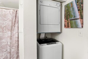 A laundry area with a front-loading washer and dryer, a shower curtain, and a painting on the wall. - Kona Mansions#B116 Mountain views, No Stairs, Heart of Town (Kailua Kona)