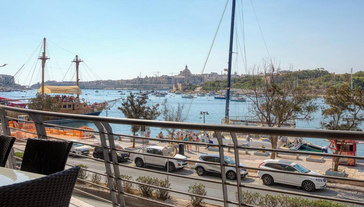 A waterfront view with a dining area overlooking the sea, boats, and a cityscape in the background.