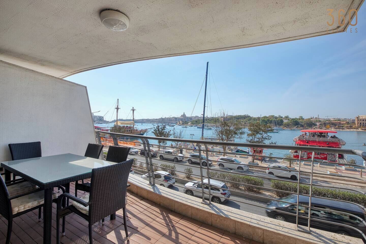 A balcony with a table and chairs, overlooking a busy harbor with boats and cars.