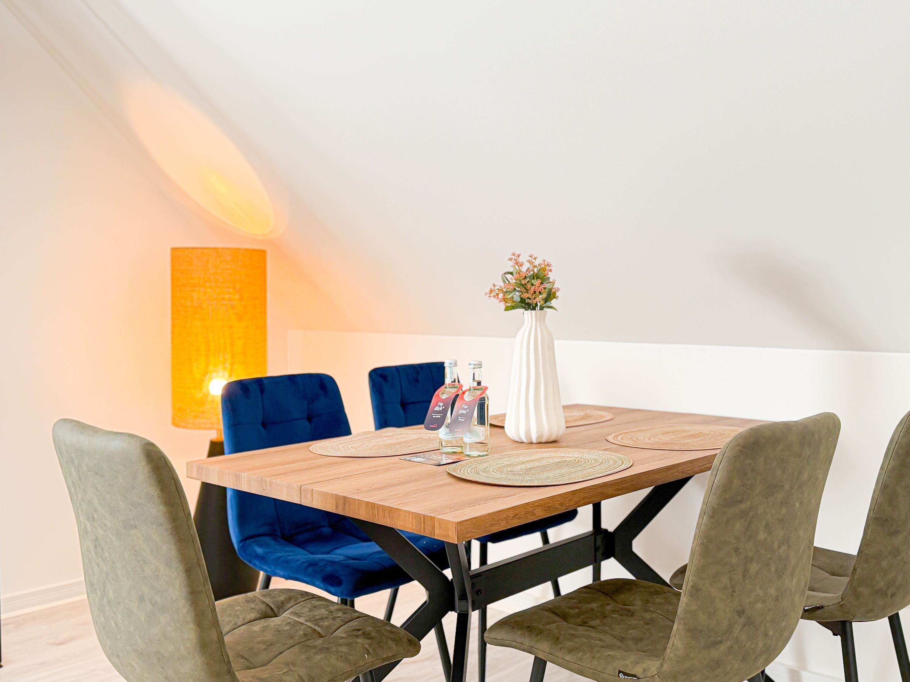 A dining area with a wooden table, blue upholstered chairs, a white vase with flowers, and a tall lamp.