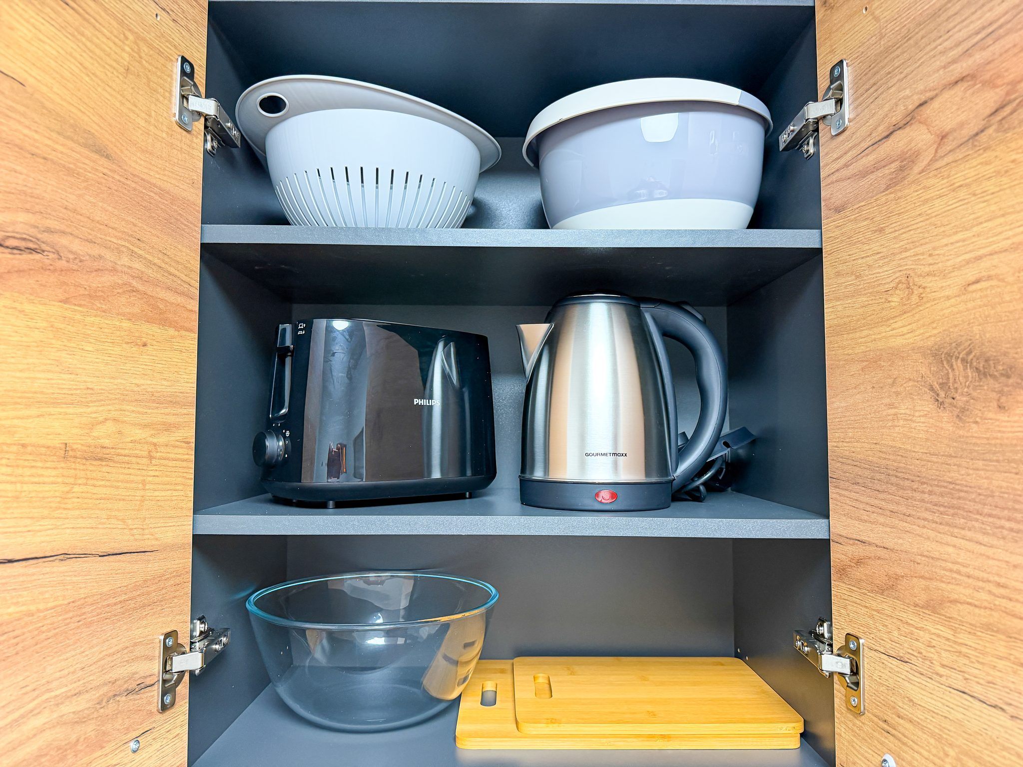 Ein Küchenschrank mit Regalen, auf denen Geschirr, darunter ein Sieb, Schüsseln, eine Kaffeekanne, ein Wasserkocher, eine Glaskugel und ein Schneidebrett, steht.
