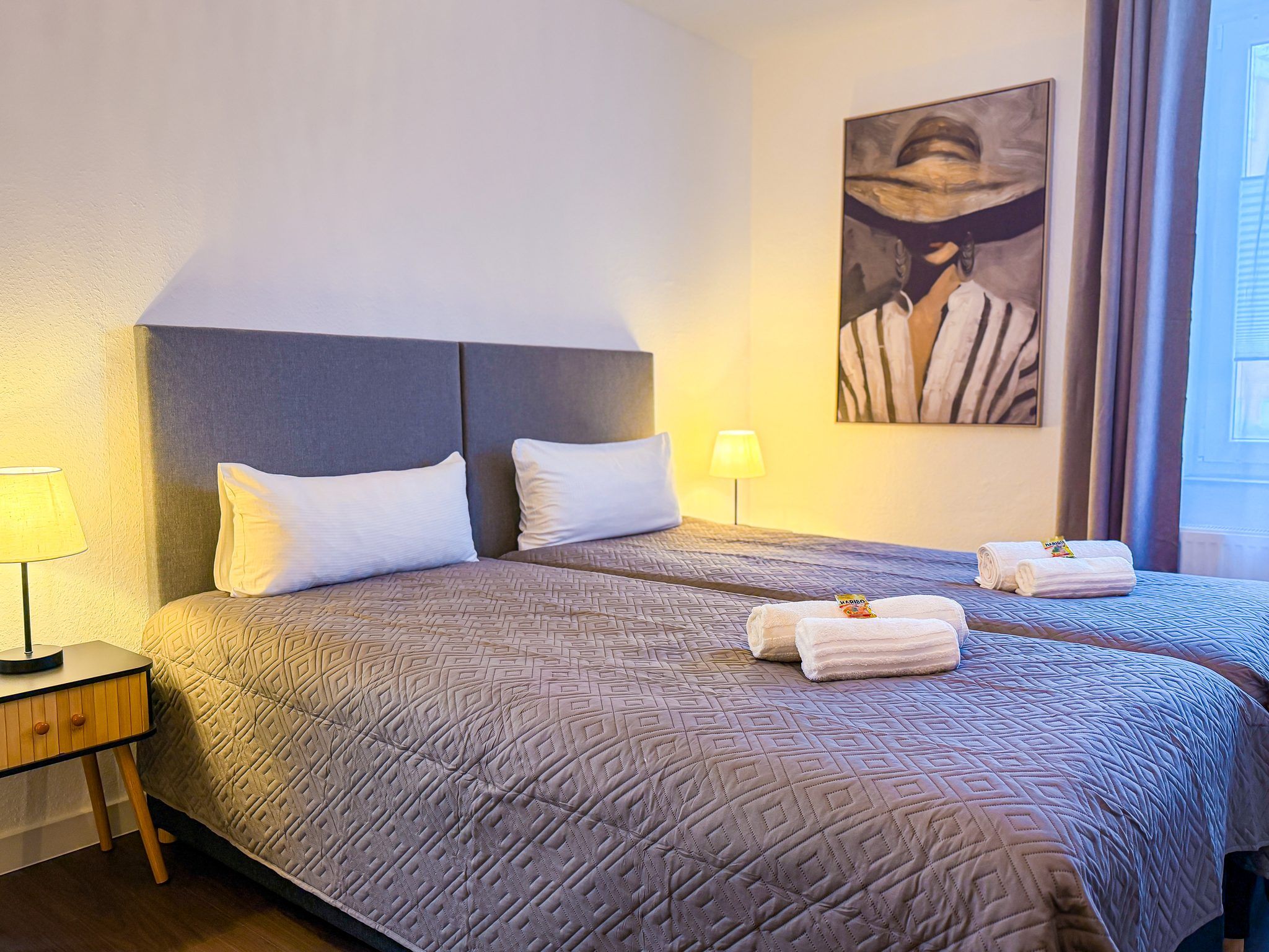 A hotel room with a large bed, two pillows, a nightstand with a lamp, and a painting on the wall.