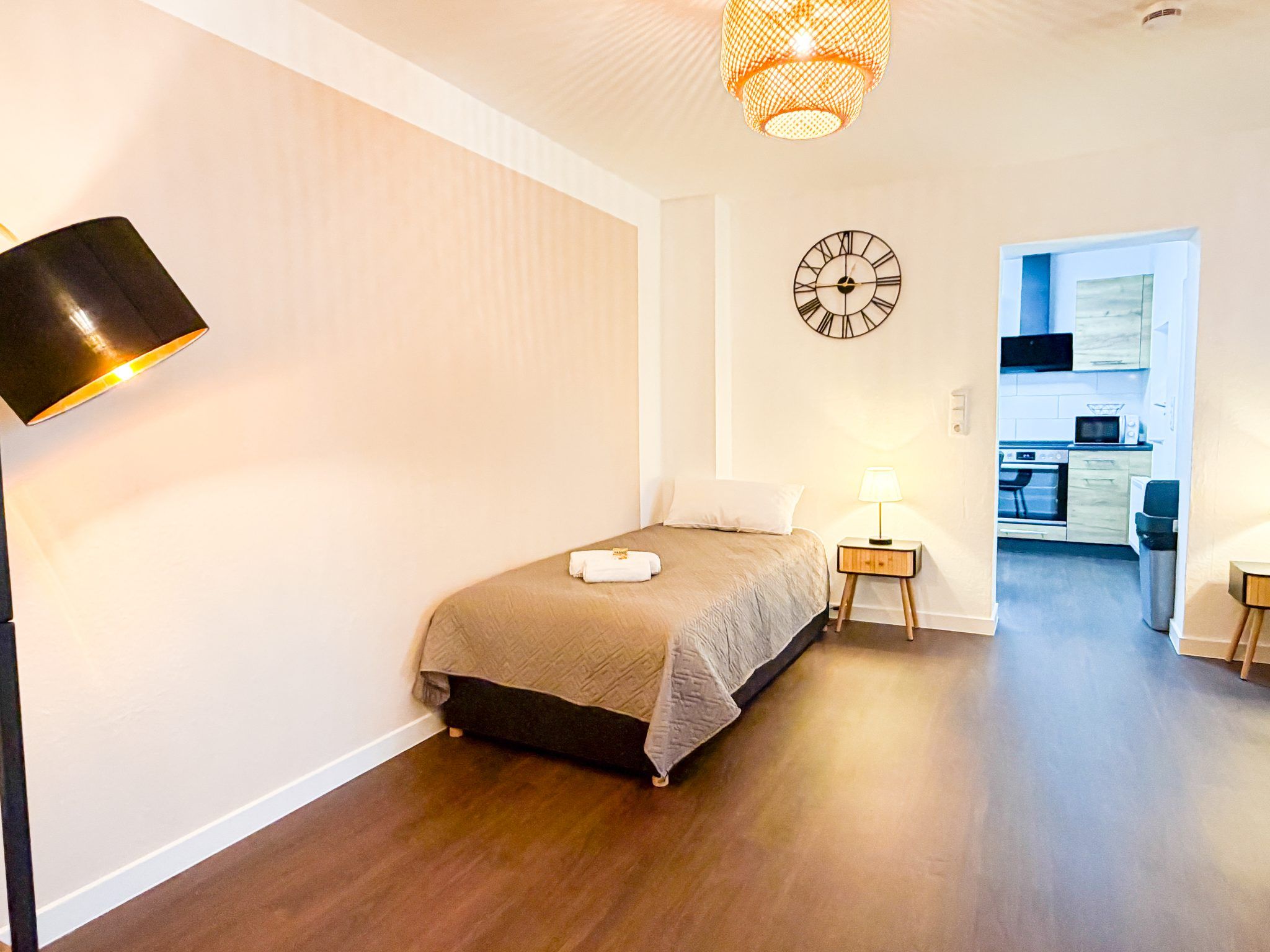 A hotel room with a bed, bedside lamps, a clock, and a view into the kitchen.