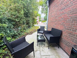 Een terras met rieten meubels en een glazen tafel, naast een bakstenen muur.