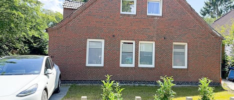 A red brick house with two windows, a car parked in front, and a chain-link fence with plants.