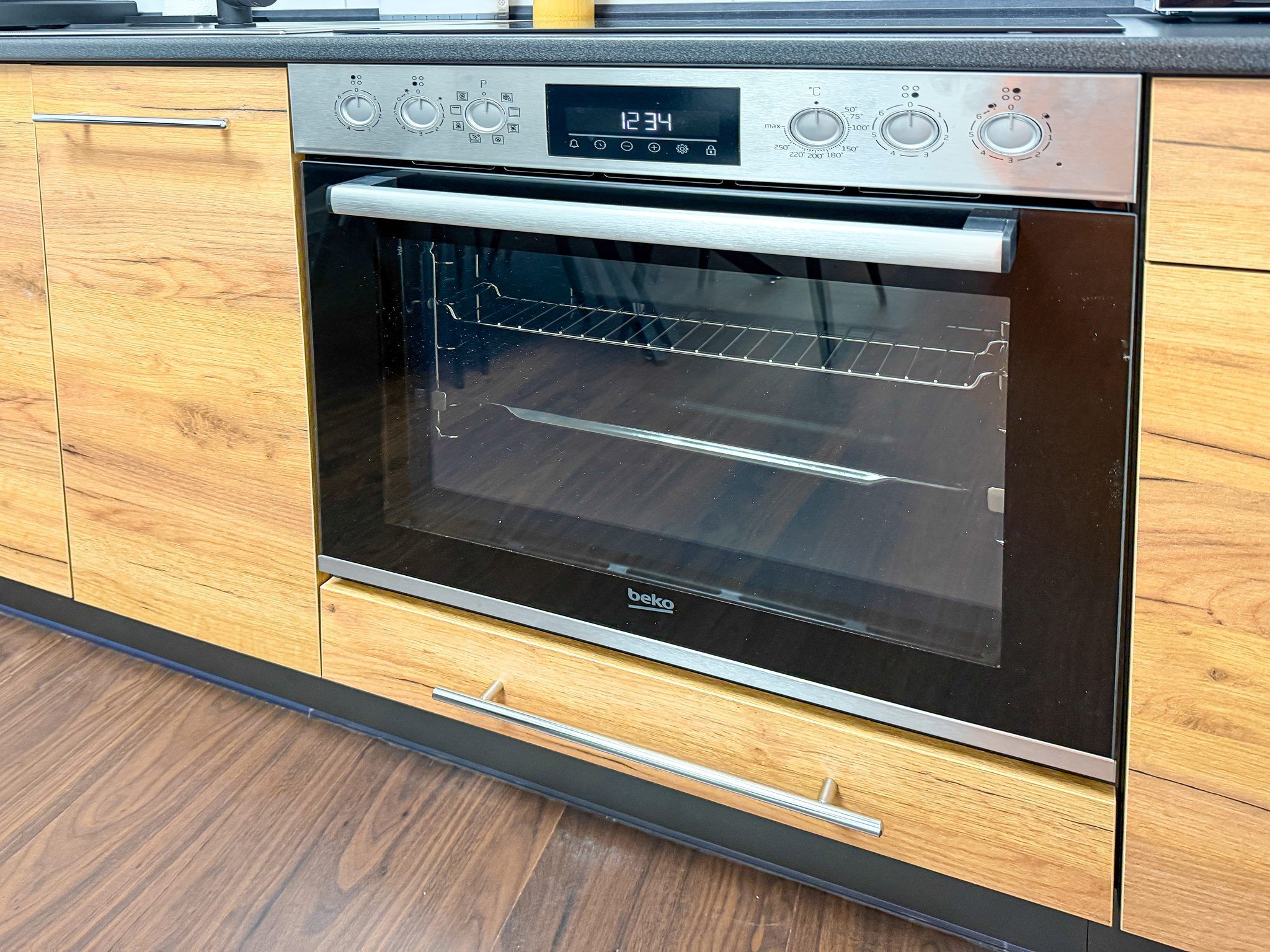 A modern kitchen with a built-in oven, wooden cabinetry, and a digital clock displaying the time.