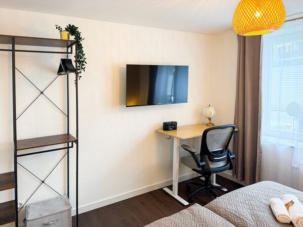 Habitación moderna con televisión en la pared, un escritorio con silla, una estantería y una cama con edredón estampado.