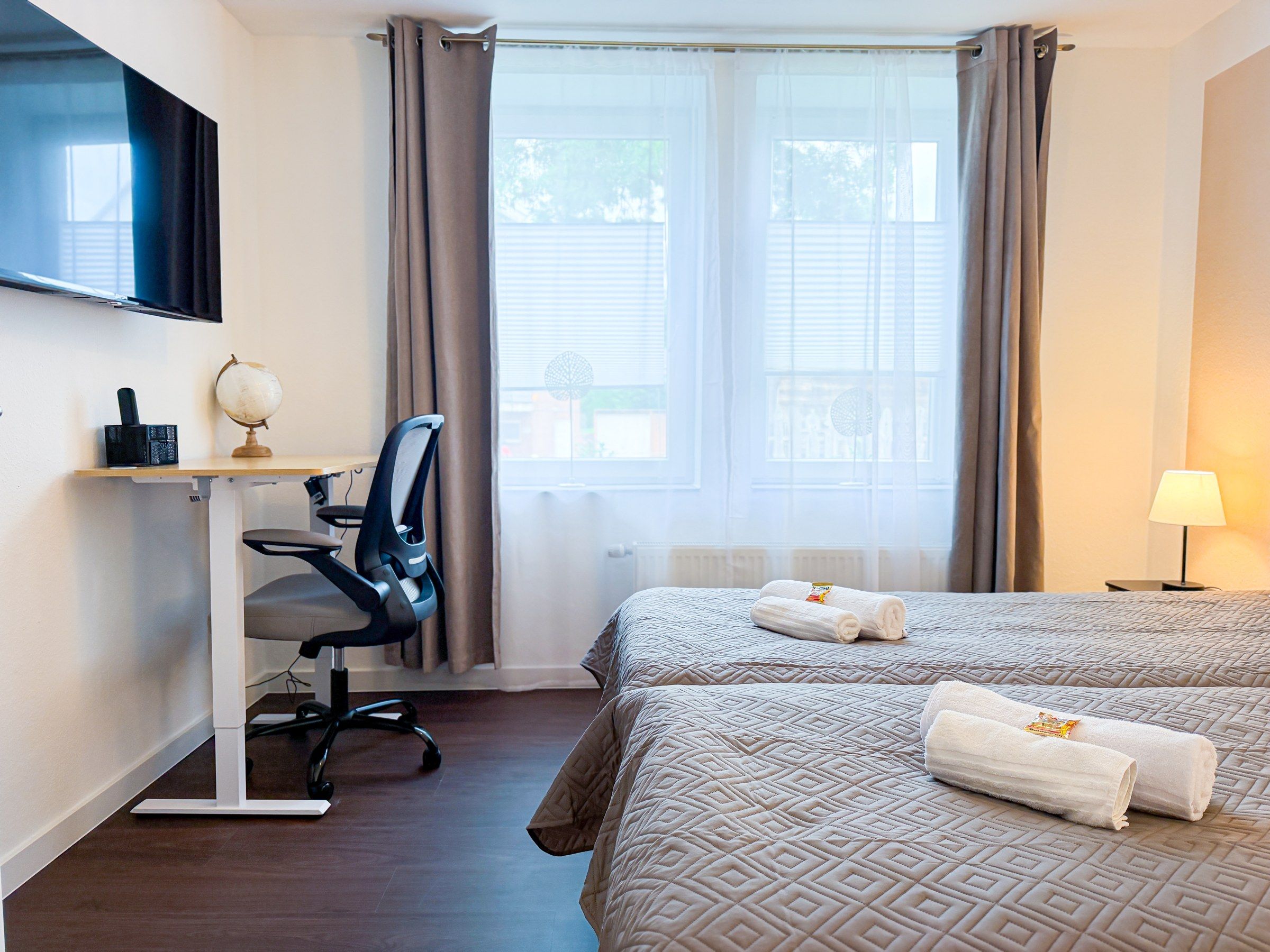 Ein Hotelzimmer mit einem Bett, einem Schreibtisch mit Stuhl, einem Fernseher und einem Fenster mit Vorhängen.
