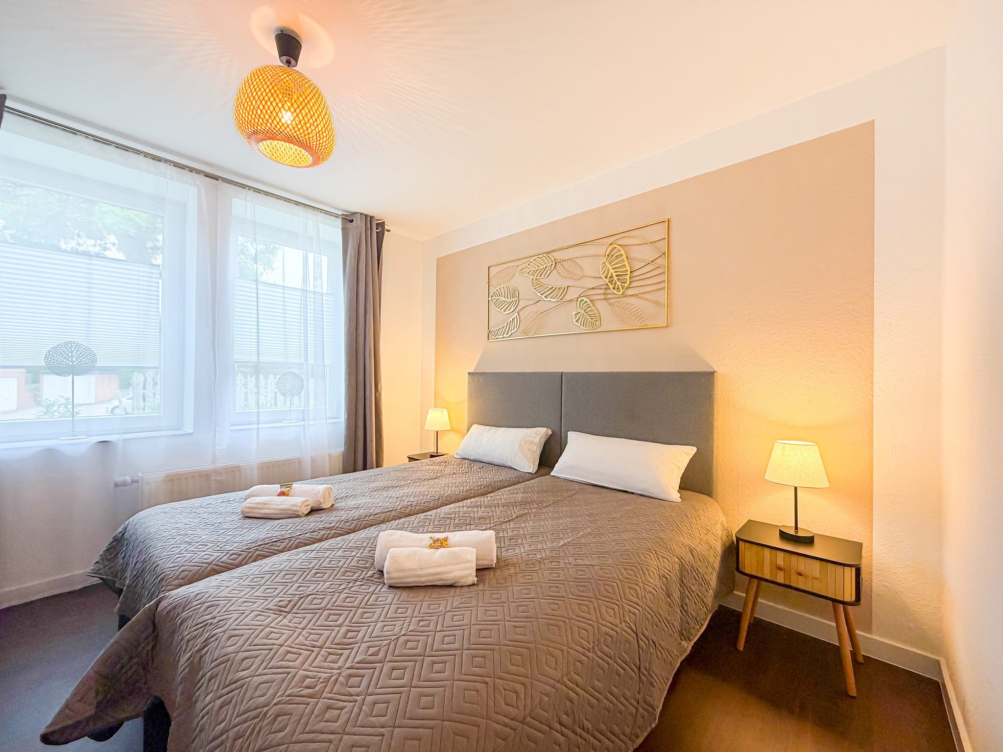 A hotel room with two beds, a window with curtains, a wall-mounted artwork, and a bedside table with a lamp.
