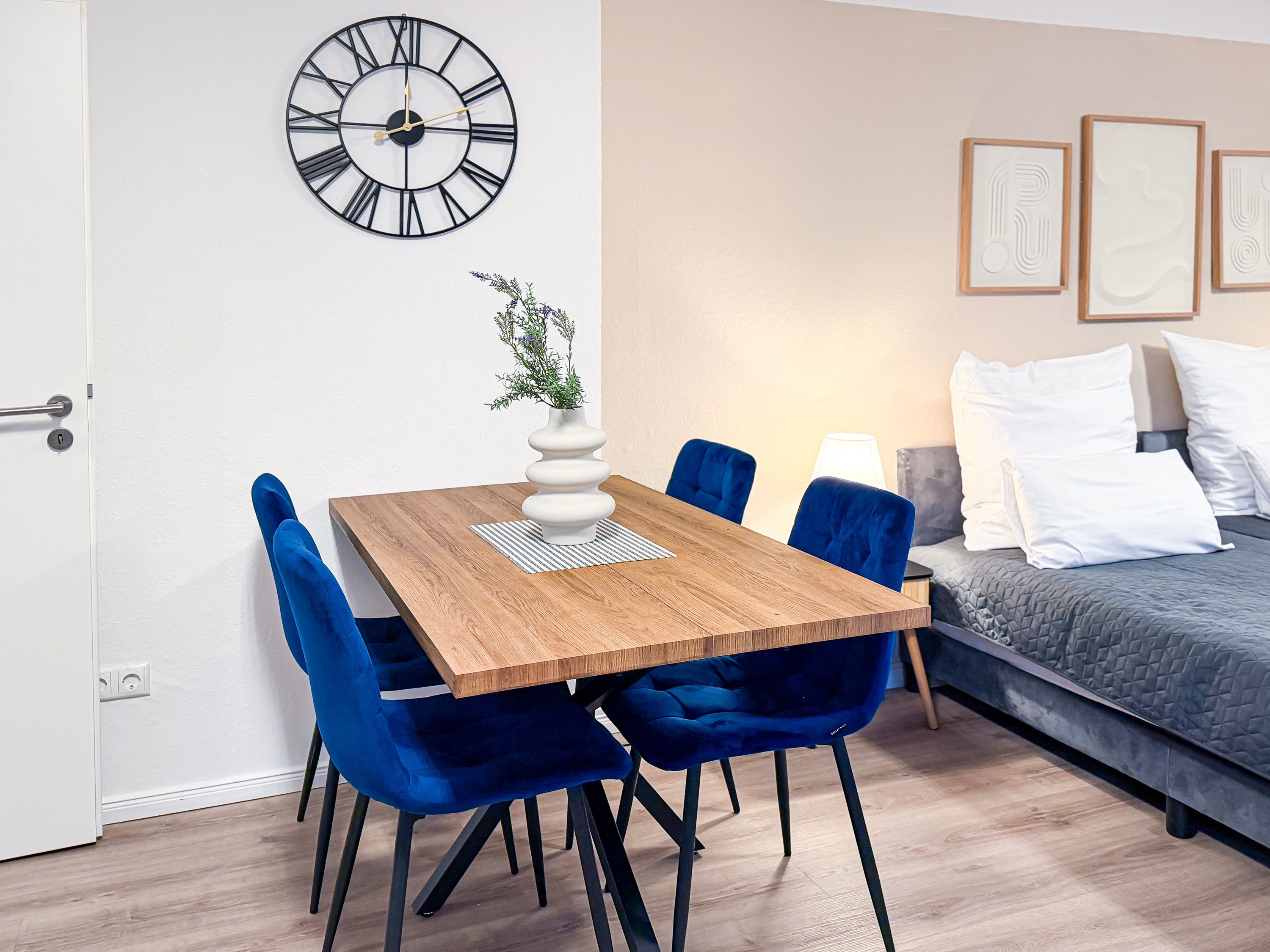 Une pièce moderne avec une table à manger en bois, des chaises bleues, une horloge, un lit avec des oreillers et des œuvres d’art encadrées accrochées au mur.