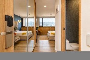 A modern hotel room with a large bed, a smaller sofa, a desk, and a view of the city through large windows. - Go Living Suites - 1101 (Medellín)