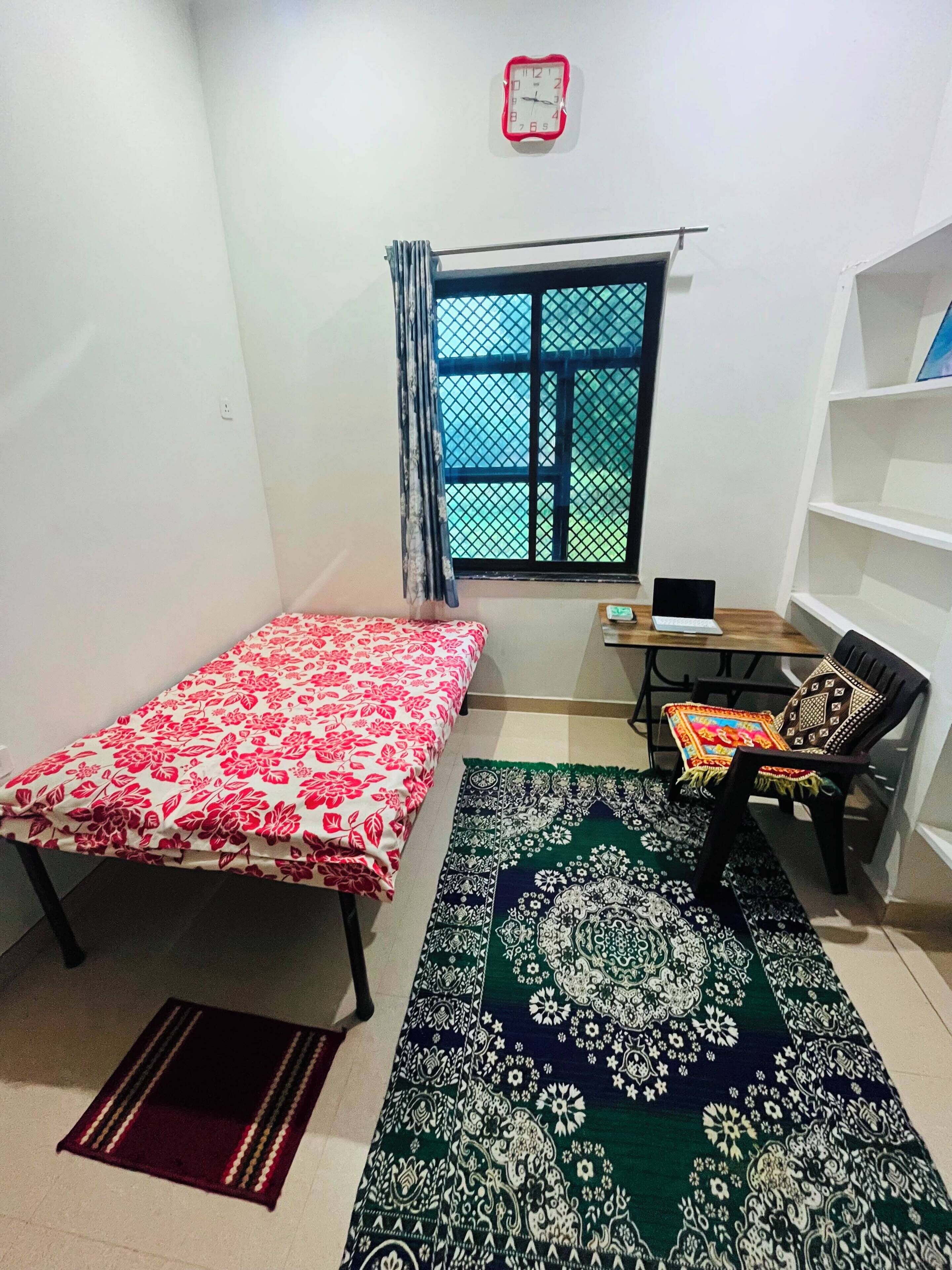 Una habitación pequeña y escasamente amoblada, con una cama, un escritorio, una silla, una ventana con reja de metal y una estantería.
