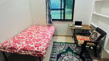 Una habitación pequeña y escasamente amoblada, con una cama, un escritorio, una silla, una ventana con reja de metal y una estantería.
