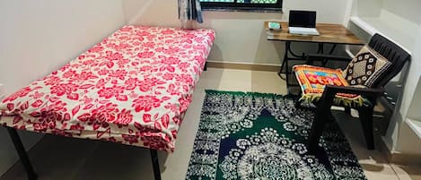 A small, sparsely furnished room with a bed, a desk, a chair, a window with a metal grill, and a bookshelf.