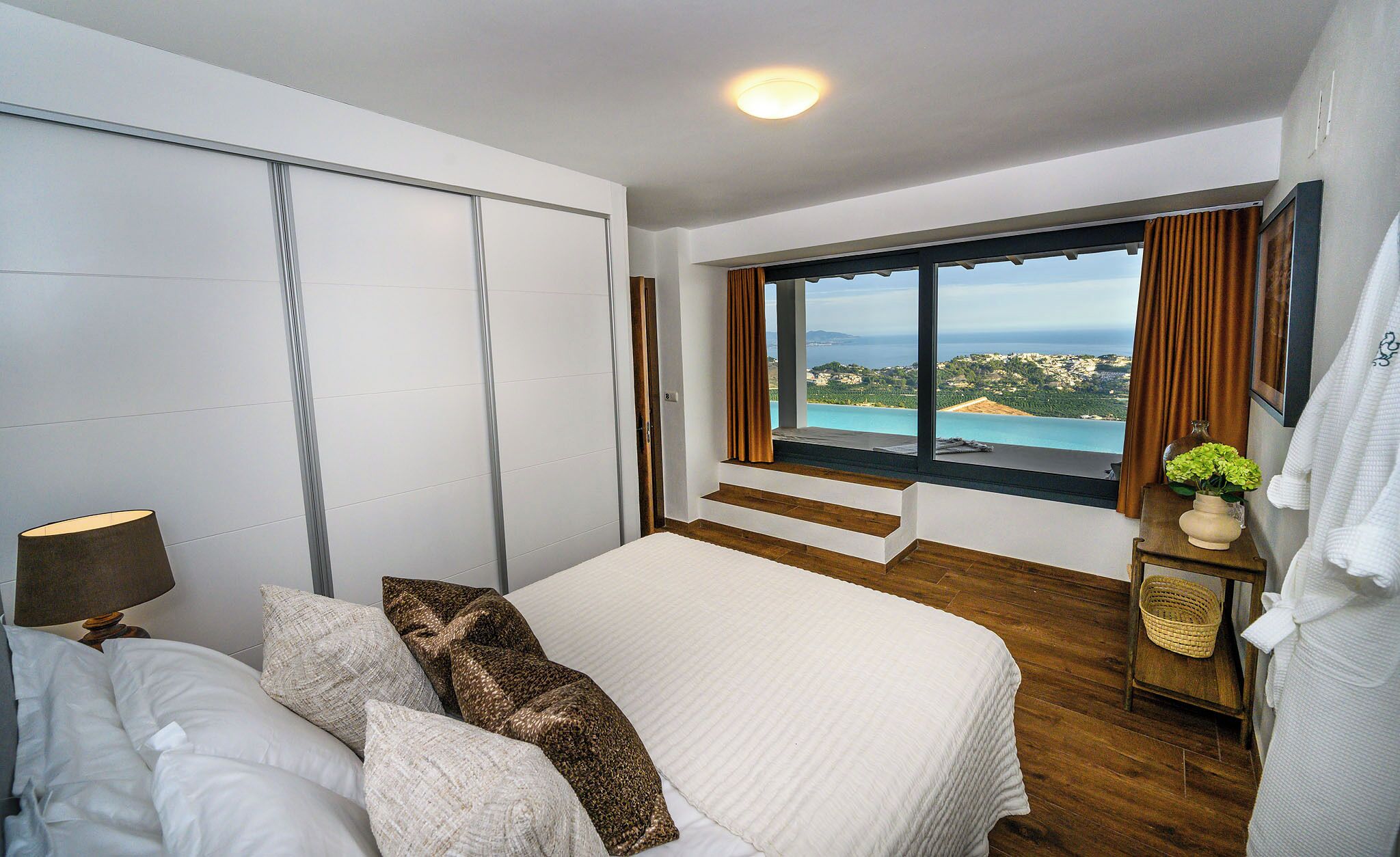 Un dormitorio con una cama grande, una mesita de noche, una lámpara, un ventanal con vista al mar y una puerta corrediza.