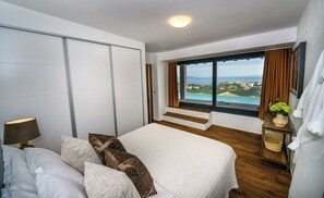 Un dormitorio con una cama grande, una mesita de noche, una lámpara, un ventanal con vista al mar y una puerta corrediza.