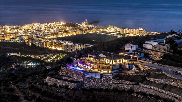 Un pueblo costero con un edificio destacado en la cima de una colina, piscina y vista al mar.