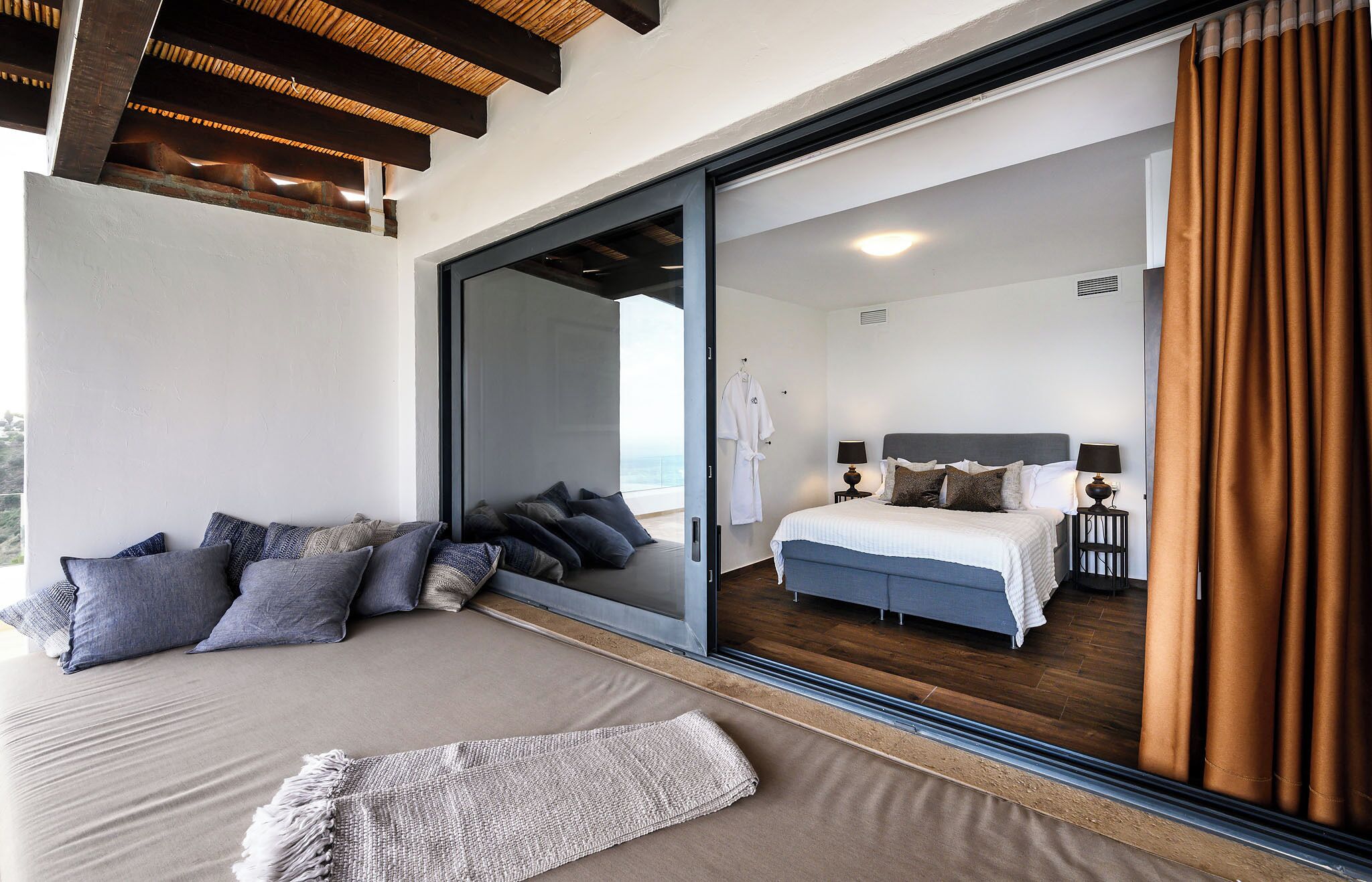 Un dormitorio con un amplio ventanal corredera que da a un balcón, una cama con cabecera gris y una mesita de noche con lámparas.