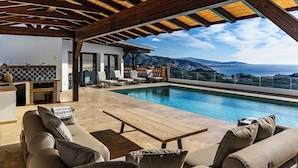 Zona junto a la piscina con una pérgola de madera, mobiliario exterior y vista al océano.