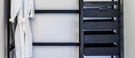 Een zwarte metalen garderobe met meerdere planken en een kledingroede, met daarin een witte badjas en een klein elektronisch apparaat.