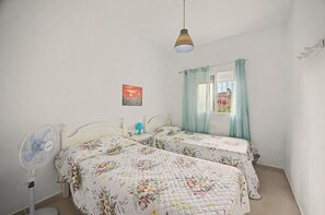 Ein Schlafzimmer mit zwei Betten, einem Nachttisch mit Lampe, einem an der Wand montierten Ventilator und einem Fenster mit Vorhängen.