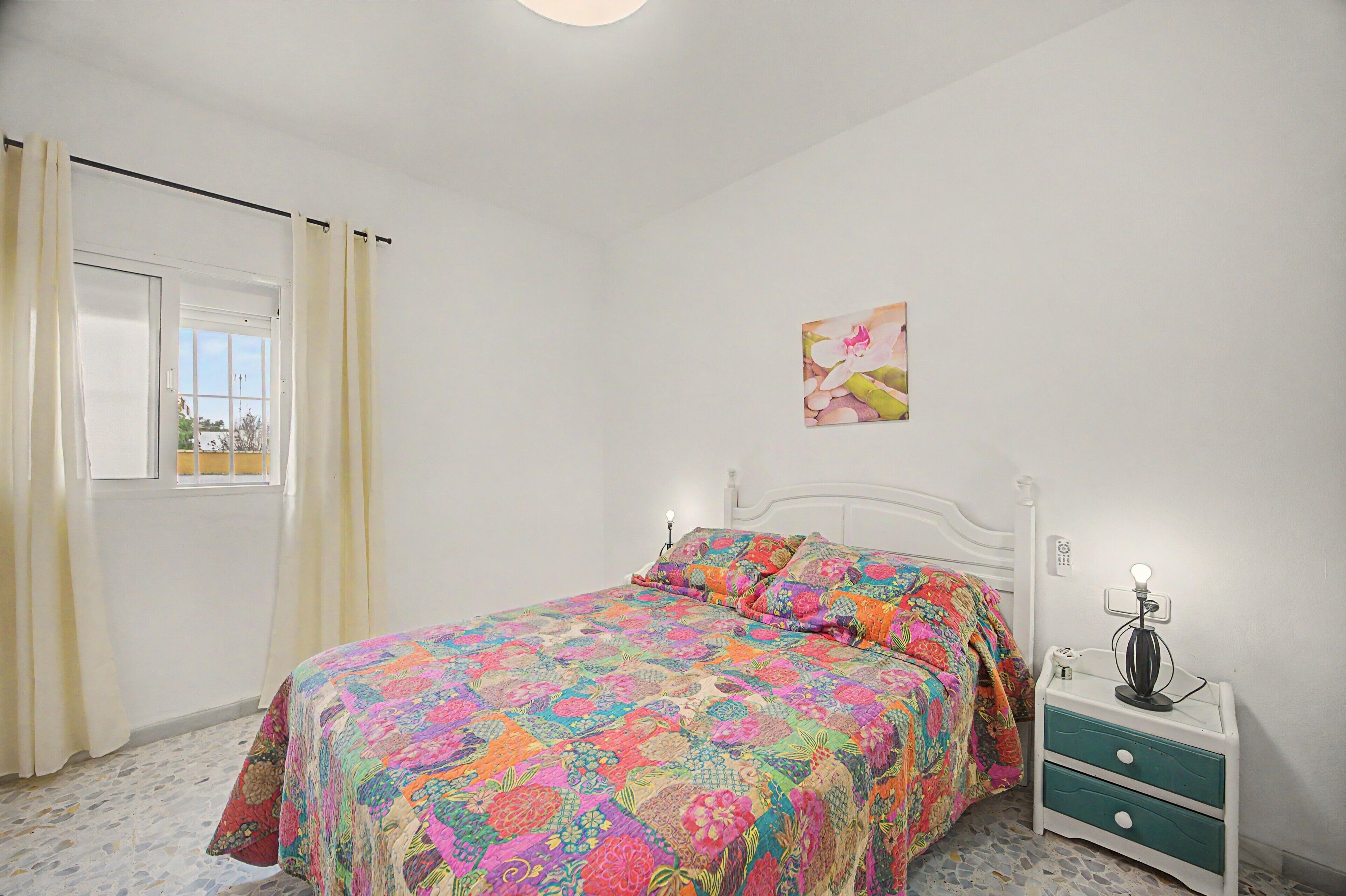 Une chambre à coucher comprenant un lit, une table de chevet, une commode, une fenêtre avec des rideaux et un luminaire fixé au mur.
