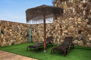 A stone wall with a woven overhead structure, two lounge chairs, and artificial grass. - Holiday Home 'La Chumbera' with Shared Pool and Private Terrace (Vejer de la Frontera)