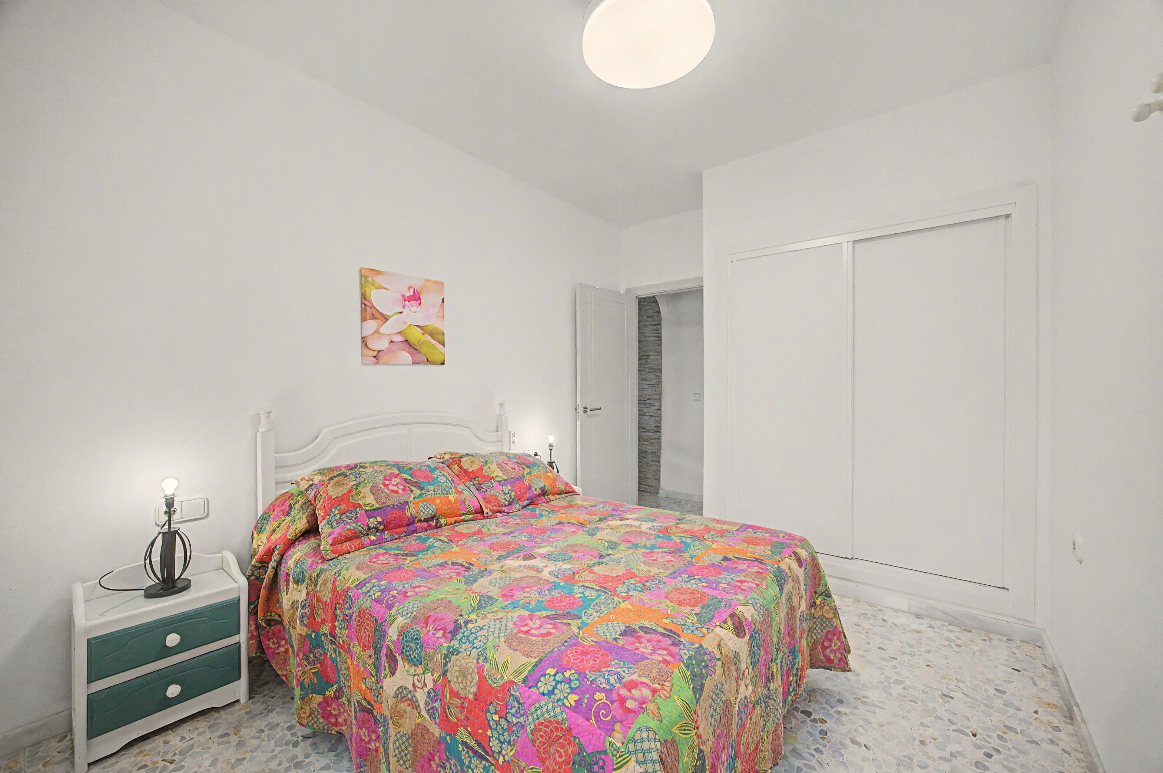 A bedroom with a bed, a nightstand, a wall-mounted light, and a door.