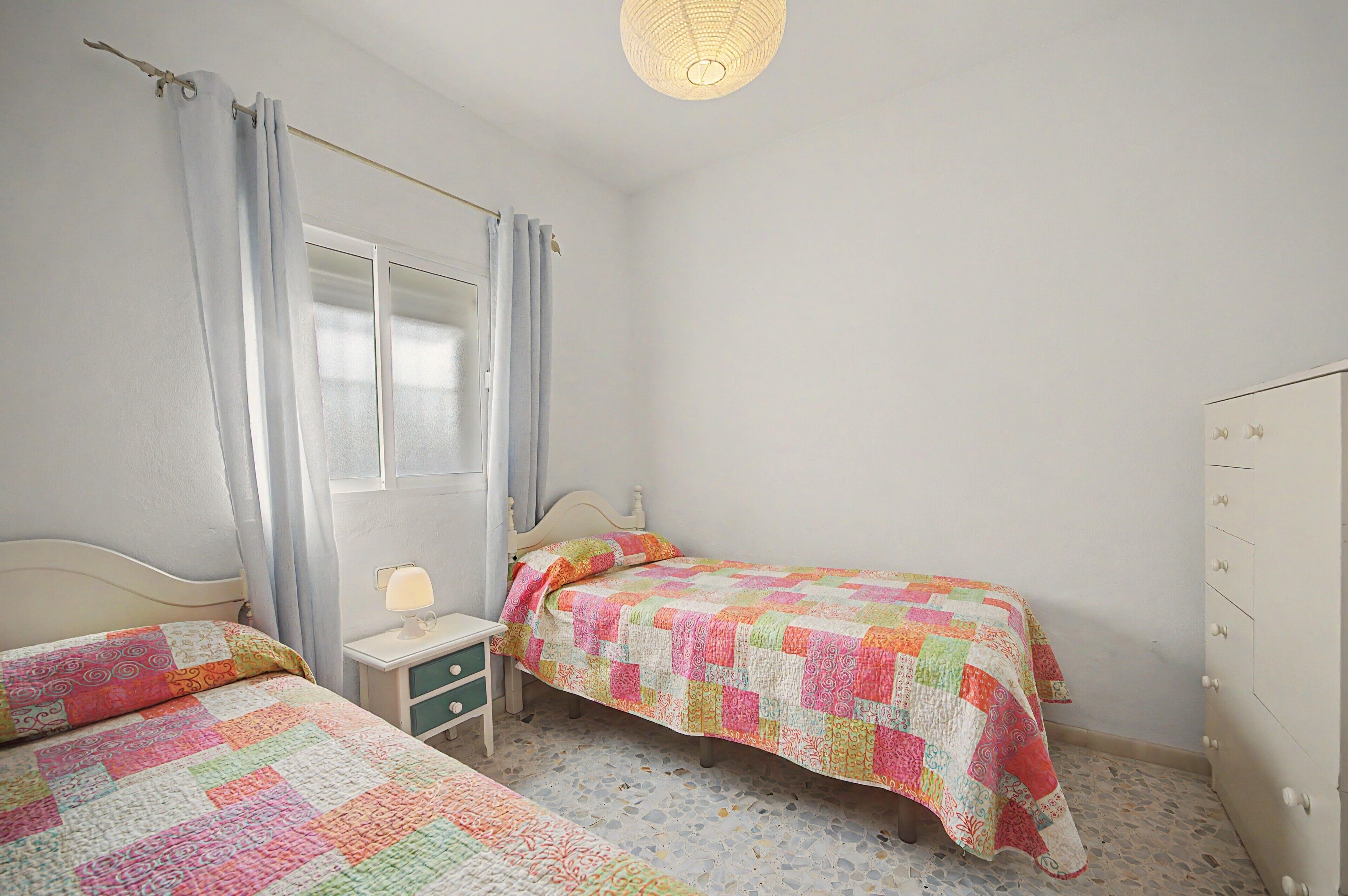 A bedroom with two beds, a window with curtains, a bedside table with a lamp, and a wardrobe.